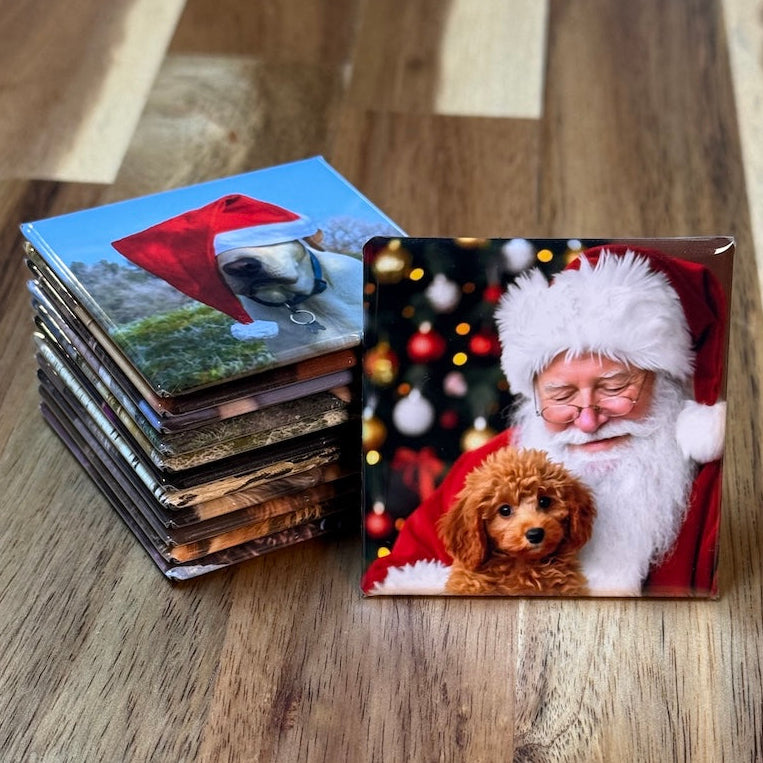 Stack of 2x2 inch custom photo magnets on a wooden table, with a Christmas magnet of Santa holding a puppy standing in front