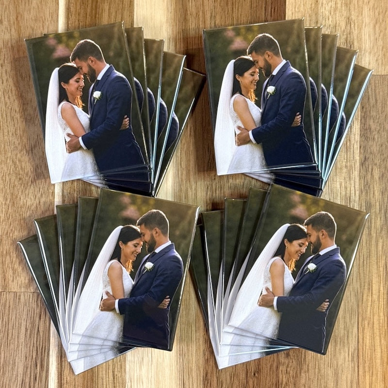 Twenty identical 2×3 inch rectangle photo magnets of a wedding couple, fanned in four stacks on a wood table, made as wedding favors